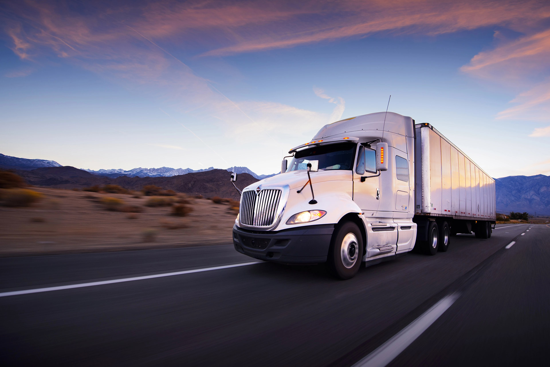 A silver semi-truck drives along a highway, surrounded by desert landscape and mountains under a colorful sunset sky.