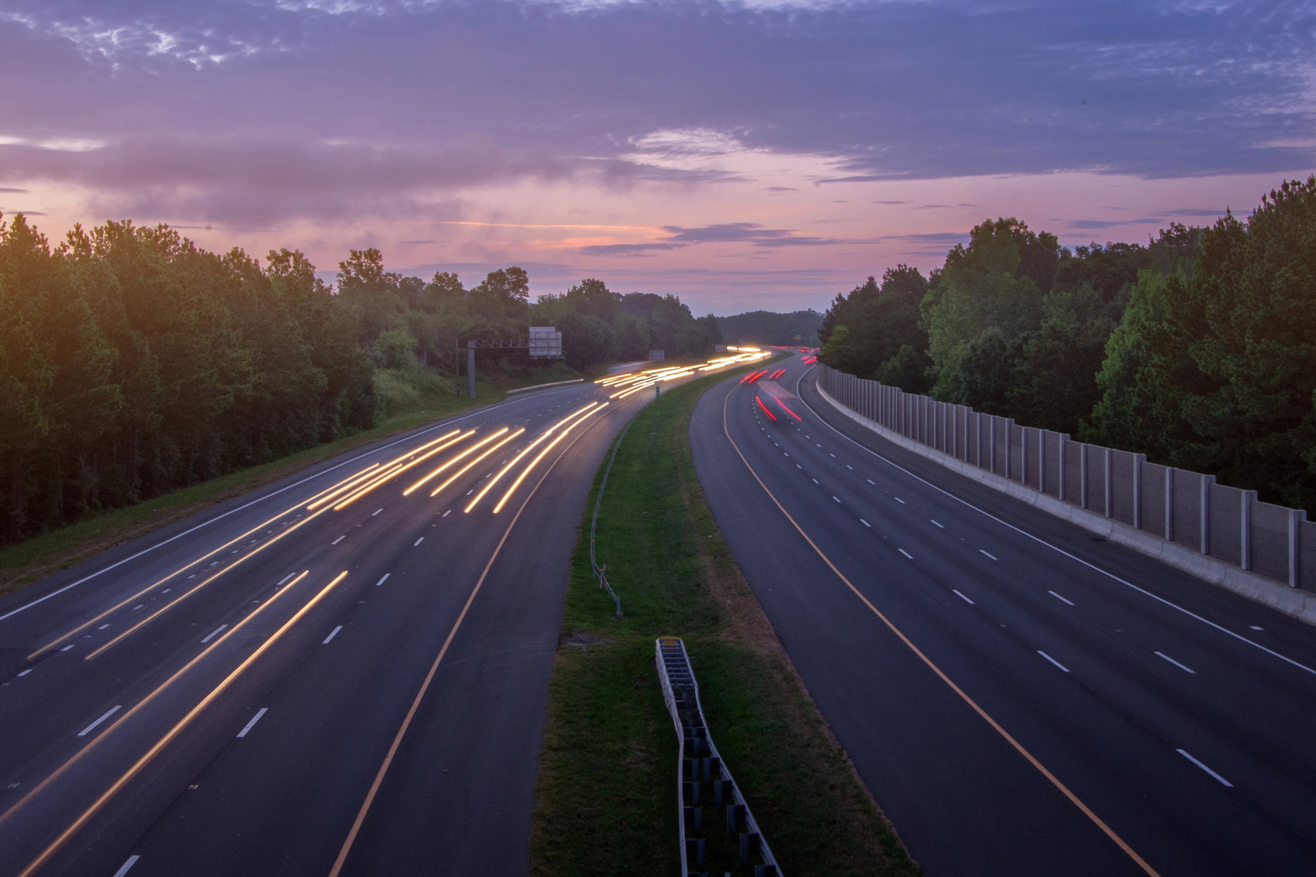 A twilight scene of a highway shows streaks of car lights, flanked by greenery and sound barriers, under a softly lit sky.