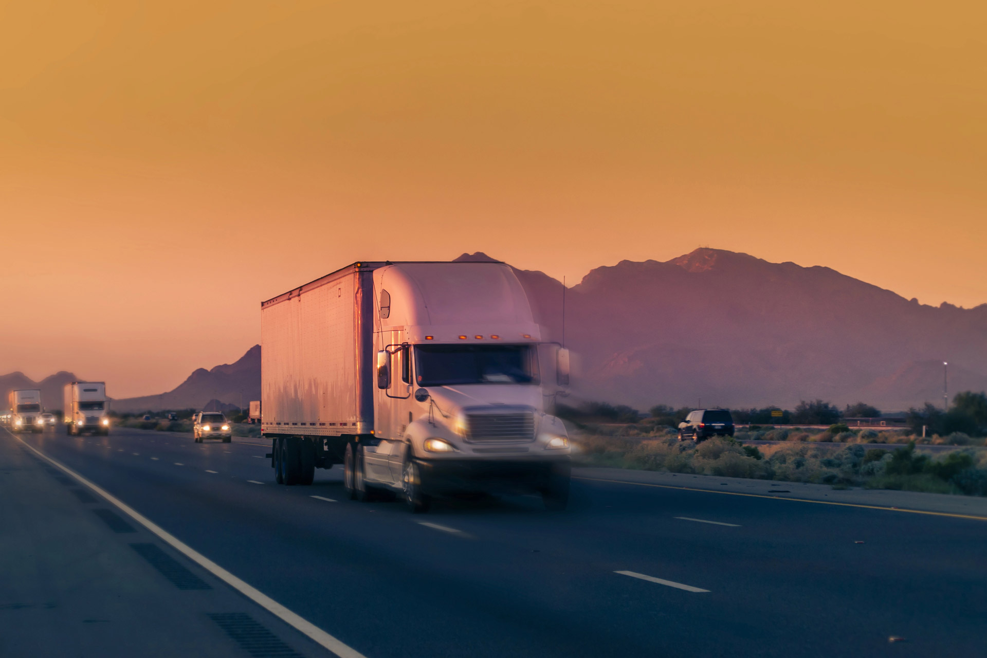 A pink-hued sunset illuminates a highway where a semi-truck speeds past mountainous scenery, creating a dynamic sense of motion.