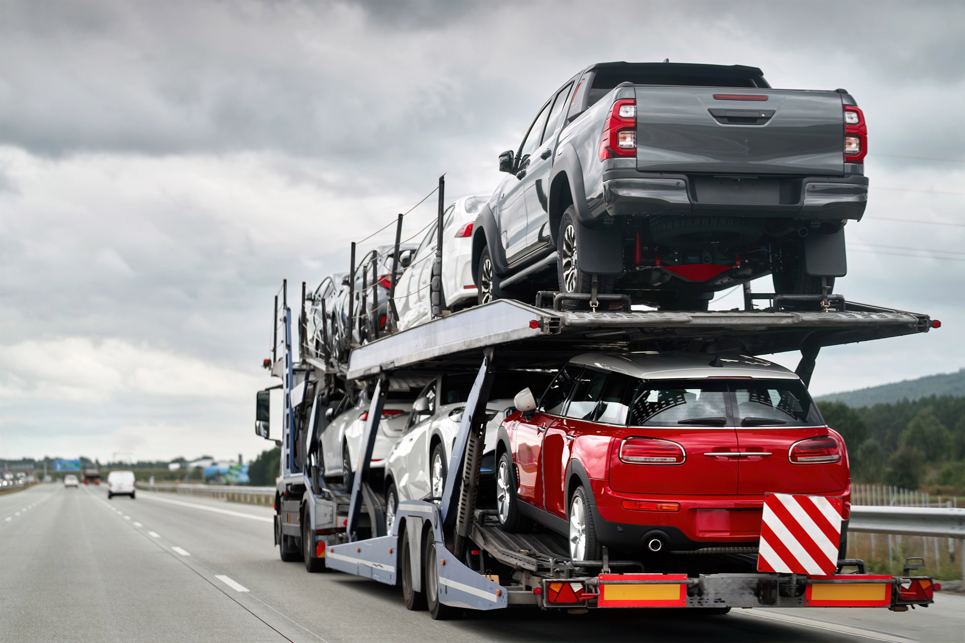 A car carrier truck transports multiple vehicles, including a red sports car and several trucks, on a highway under a cloudy sky.