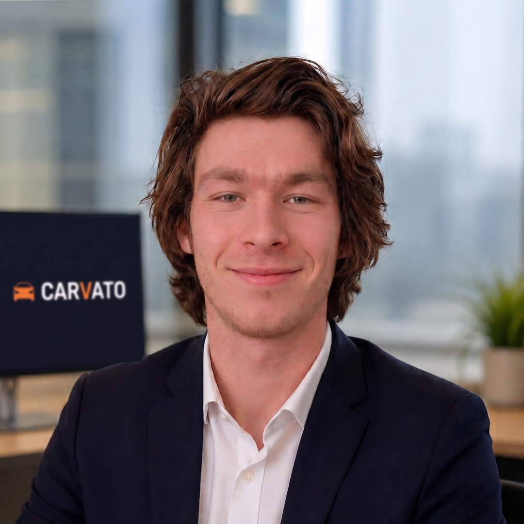 A young man with wavy hair smiles confidently, wearing a blazer and white shirt, in an office setting with a branded sign in the background.